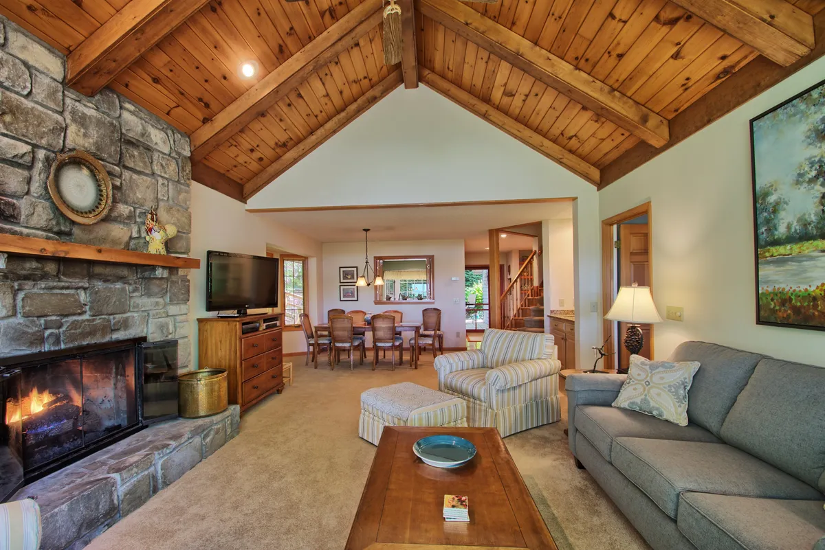 A spacious vaulted great room with exposed wooden beams, a dramatic stone fireplace as the focal point, and plush seating arranged around the fire. The layout seamlessly connects to an adjacent dining area, visible in the background, all illuminated by large windows that usher in mountain views and natural light.