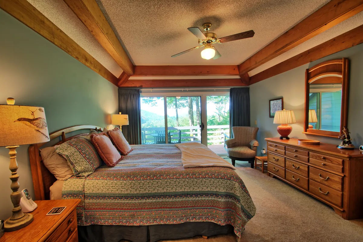 A serene bedroom with a bed adorned with decorative pillows, a large window offering a scenic outdoor view, and a wooden dresser and bedside lamp. The space feels airy, comfortable, and ideal for peaceful relaxation.