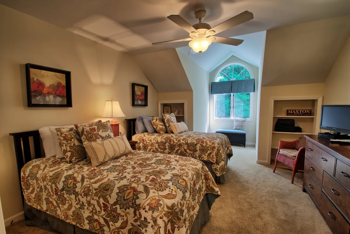 A welcoming upstairs bedroom showcasing two twin beds with decorative bedding, a window allowing in daylight, a ceiling fan, and cheerful artwork. The arrangement is ideal for children or friends traveling together, offering both comfort and a sense of personal space.
