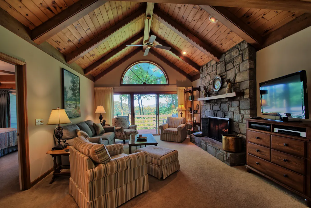 The image showcases a warm and inviting living room featuring a striking stone fireplace as the focal point, with exposed wooden beams adding rustic charm. Sunlight pours through a large window offering mountain views, while plush seating invites guests to relax, socialize, or unwind with a good book.