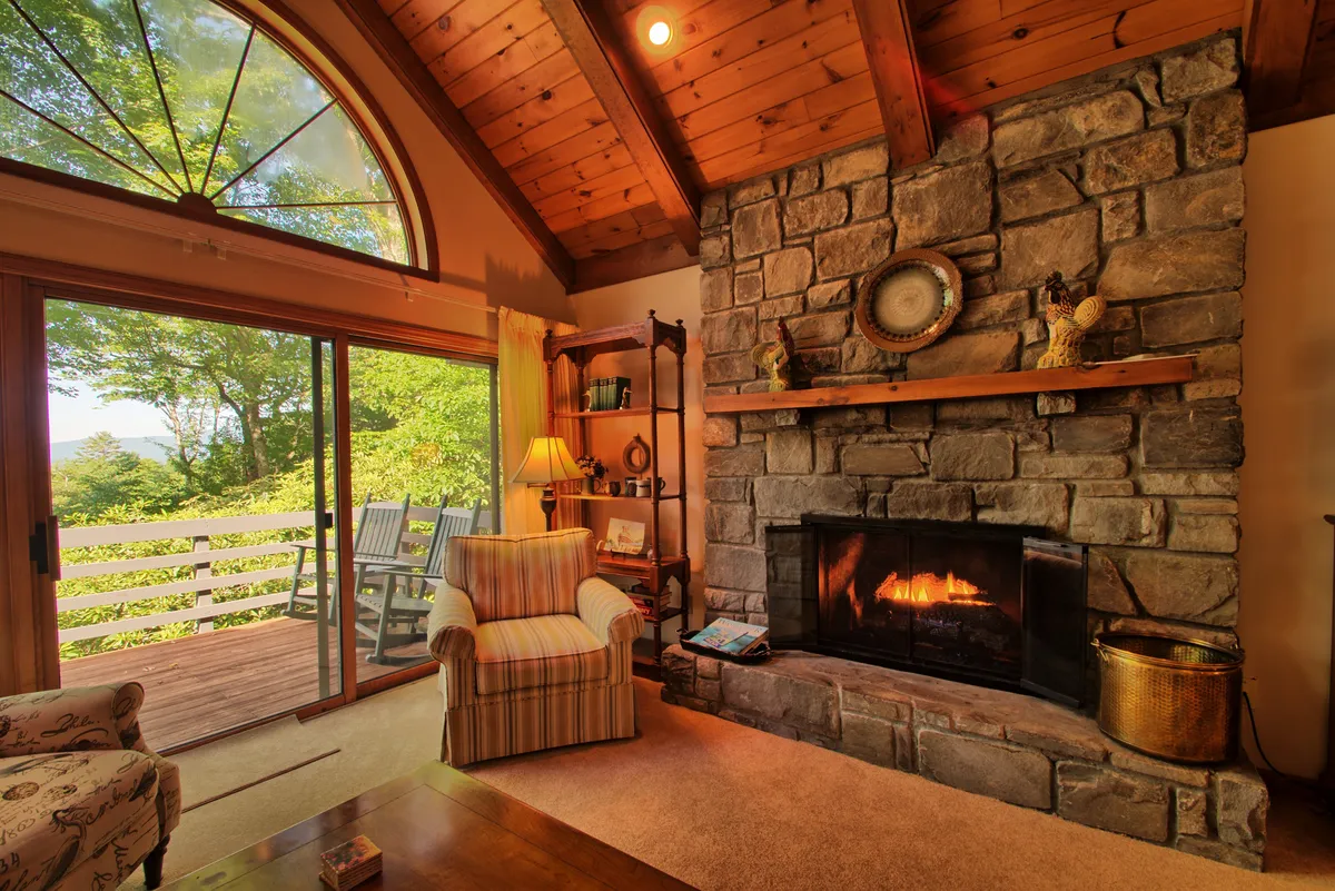 A cozy living area featuring a rustic stone fireplace, a comfortable armchair, and large windows framing tranquil mountain views. Warm sunlight filters through, creating an inviting retreat perfect for relaxing after a day’s adventures.