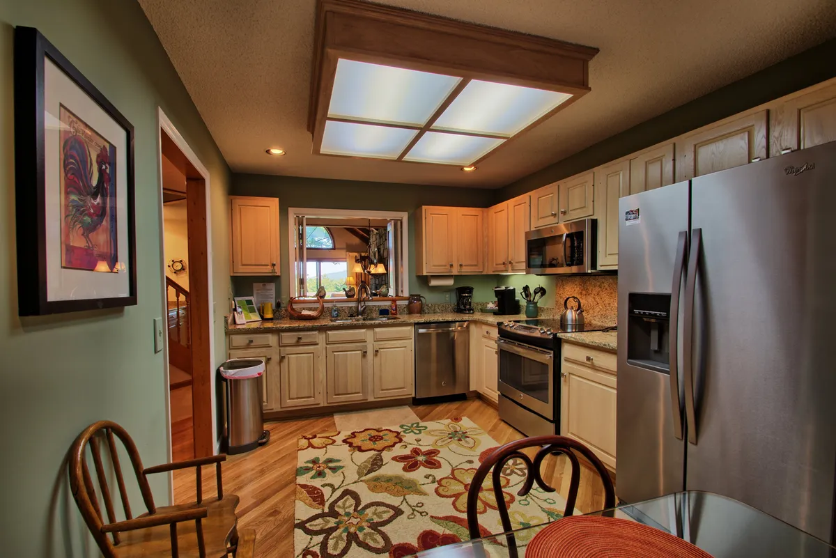 An inviting kitchen centered around a functional island, complemented by stainless steel appliances and a colorful area rug. The window in the background offers a view outside, while the layout invites both cooking and conversation.
