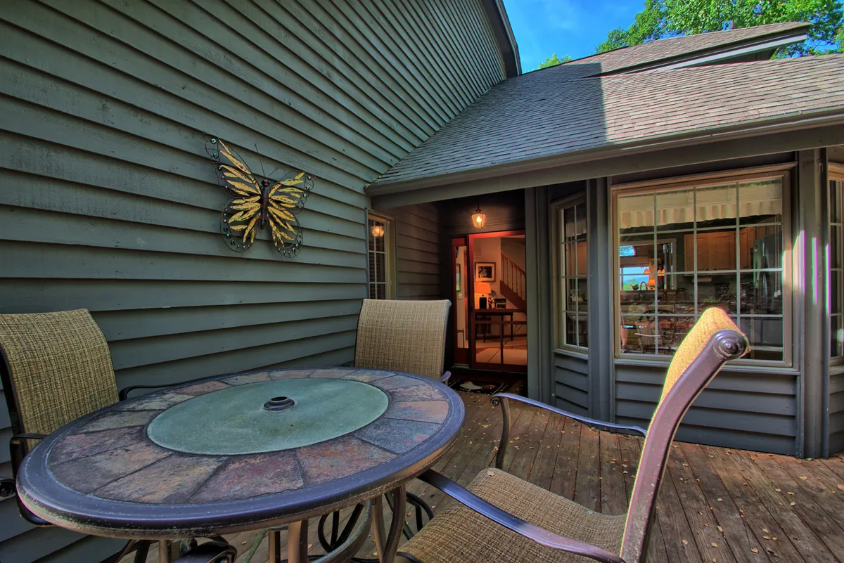A cozy exterior patio featuring a stone-topped table, comfortable chairs, and butterfly wall art. The arrangement suggests a friendly, welcoming space for outdoor dining or unwinding in the fresh air, surrounded by lush greenery.