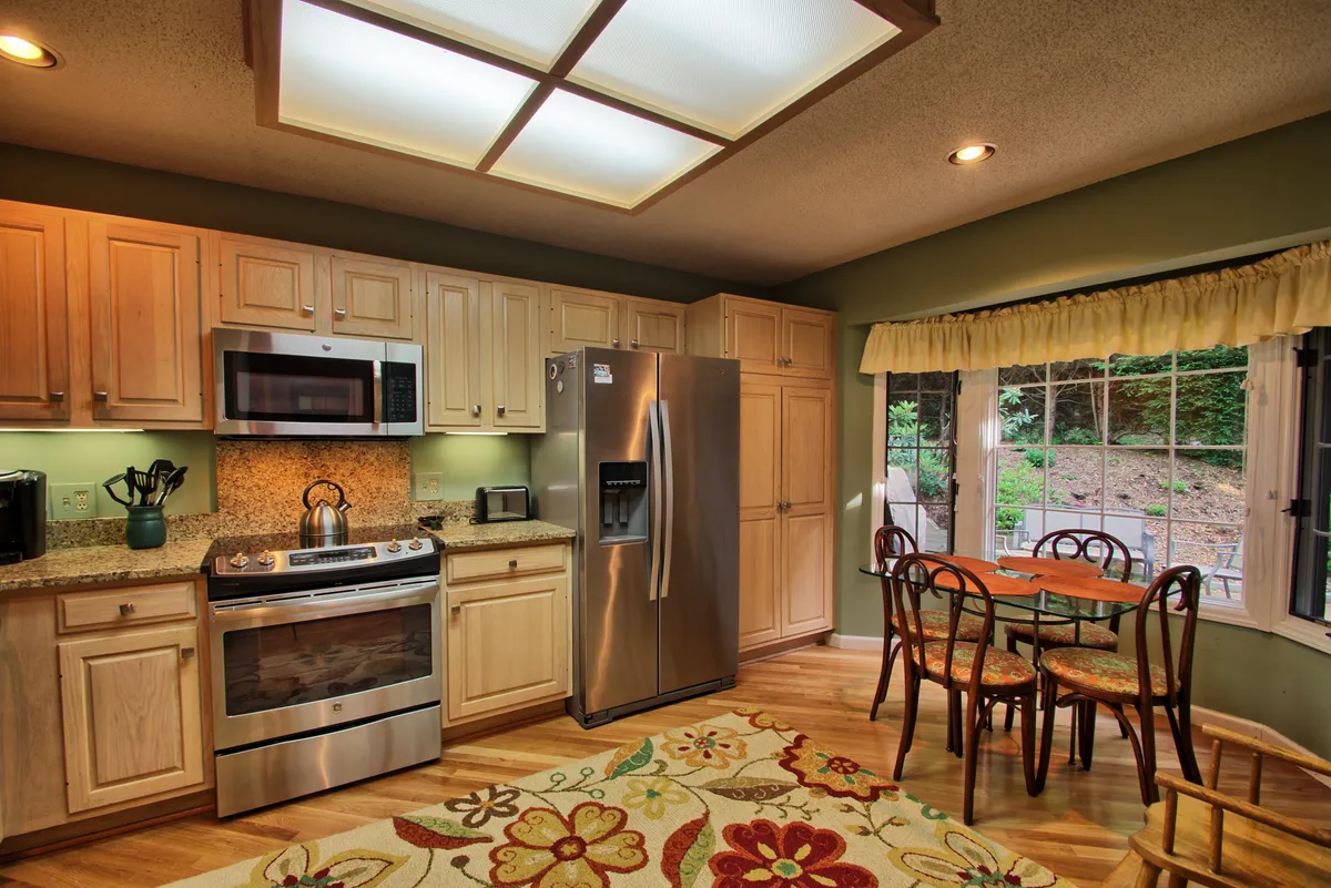 A bright, welcoming kitchen featuring white cabinetry, sleek stainless steel appliances, and a spacious dining table beside a large window with a mountain view. The space feels open and airy, with ample natural light streaming in, and is clearly designed for both cooking and gathering.