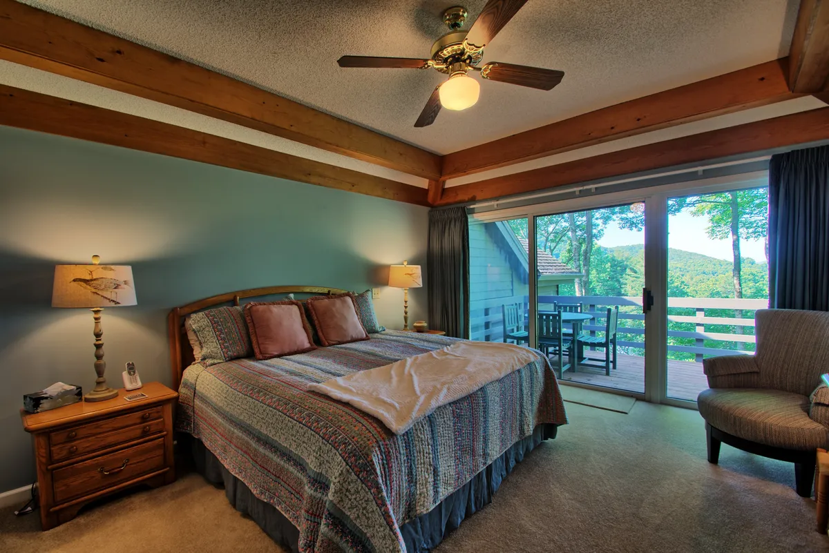 A tranquil bedroom featuring a comfortable bed positioned near a sliding glass door, with an armchair and nightstand completing the cozy arrangement. Natural light streams in through the door, which opens directly to the deck, hinting at the master suite’s private outdoor access.