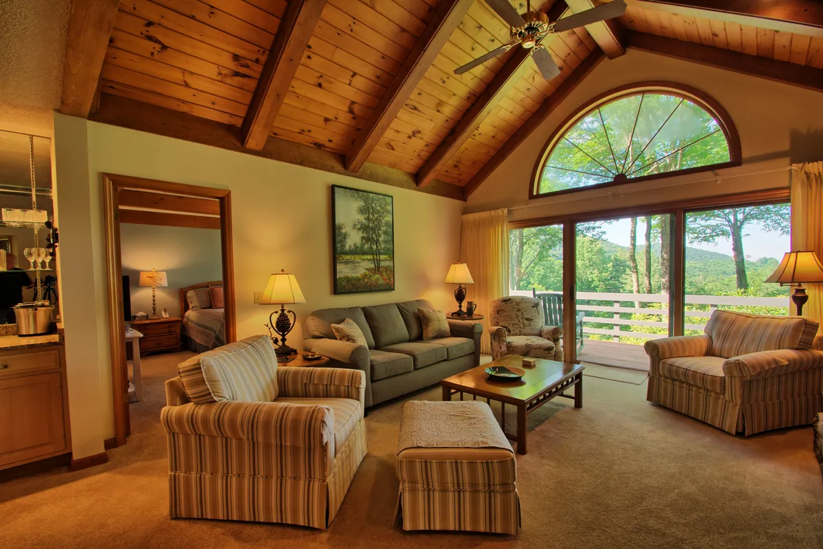 A cozy living space with comfortable sofas positioned for conversation and relaxation, highlighted by wooden ceiling beams and expansive windows framing stunning natural views. A central coffee table and casual decor reinforce a welcoming, communal atmosphere.